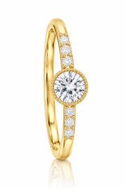 A yellow gold ring featuring a round-cut diamond center stone in a beaded halo, with smaller diamonds set along the band, shown on a clean white background with a soft reflection.