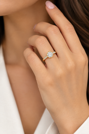 A close-up lifestyle shot of a woman's hand wearing a yellow gold ring with a round-cut clear gemstone and small side stones, set against her neck and white clothing.