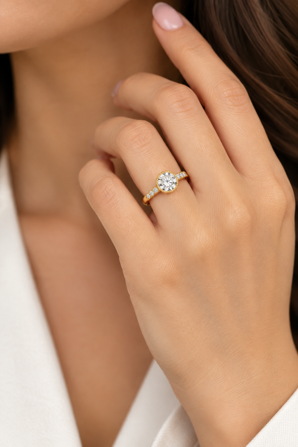 A close-up lifestyle shot of a woman's hand wearing a yellow gold ring with a round-cut clear gemstone and small side stones, set against her neck and white clothing.