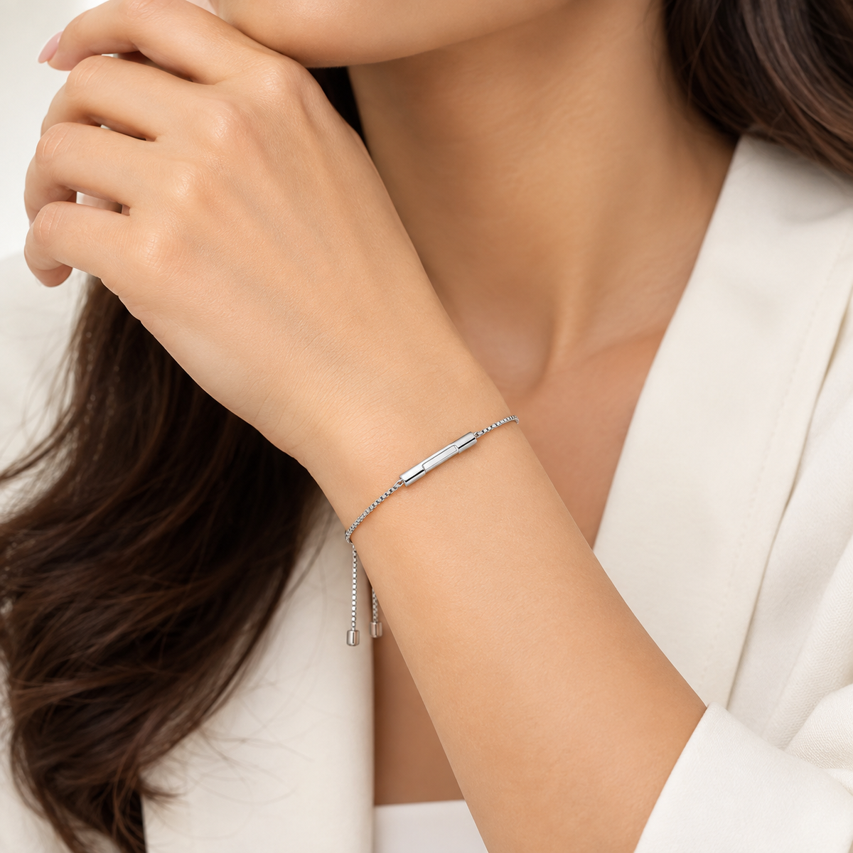 A close-up of a woman's wrist wearing a delicate silver chain bracelet with a polished bar and small dangling beads, paired with a white blazer.