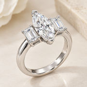 A silver-tone ring featuring a large marquise-cut clear center stone with two baguette-cut side stones, photographed on a soft cream surface with a blurred floral background.