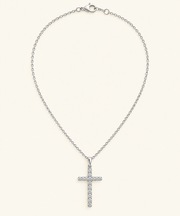A silver cross pendant necklace adorned with small clear stones, suspended from a delicate silver cable chain with a lobster clasp, set against a solid cream background.