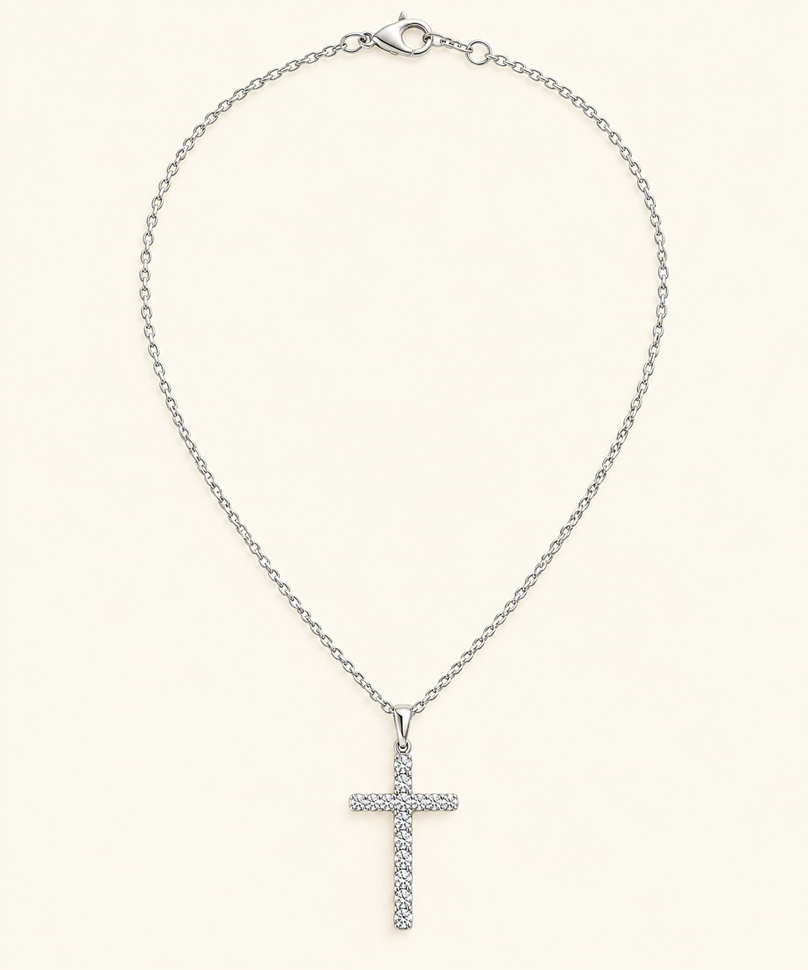A silver cross pendant necklace adorned with small clear stones, suspended from a delicate silver cable chain with a lobster clasp, set against a solid cream background.