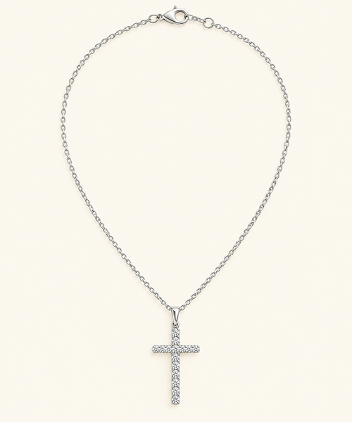 A silver cross pendant necklace adorned with small clear stones, suspended from a delicate silver cable chain with a lobster clasp, set against a solid cream background.