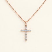 A rose gold cross pendant necklace with a pavé diamond-encrusted cross pendant on a delicate cable chain, set against a solid cream background.