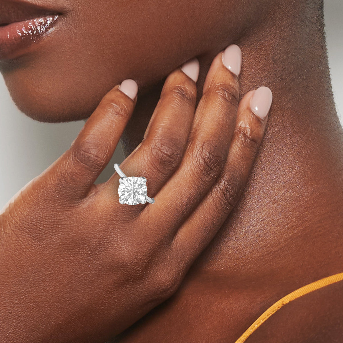 A close-up lifestyle shot of a woman's hand resting on her neck, wearing a silver engagement ring with a cushion-cut diamond. The ring is the product being sold, and the background is the model's skin and a soft neutral gray backdrop.
